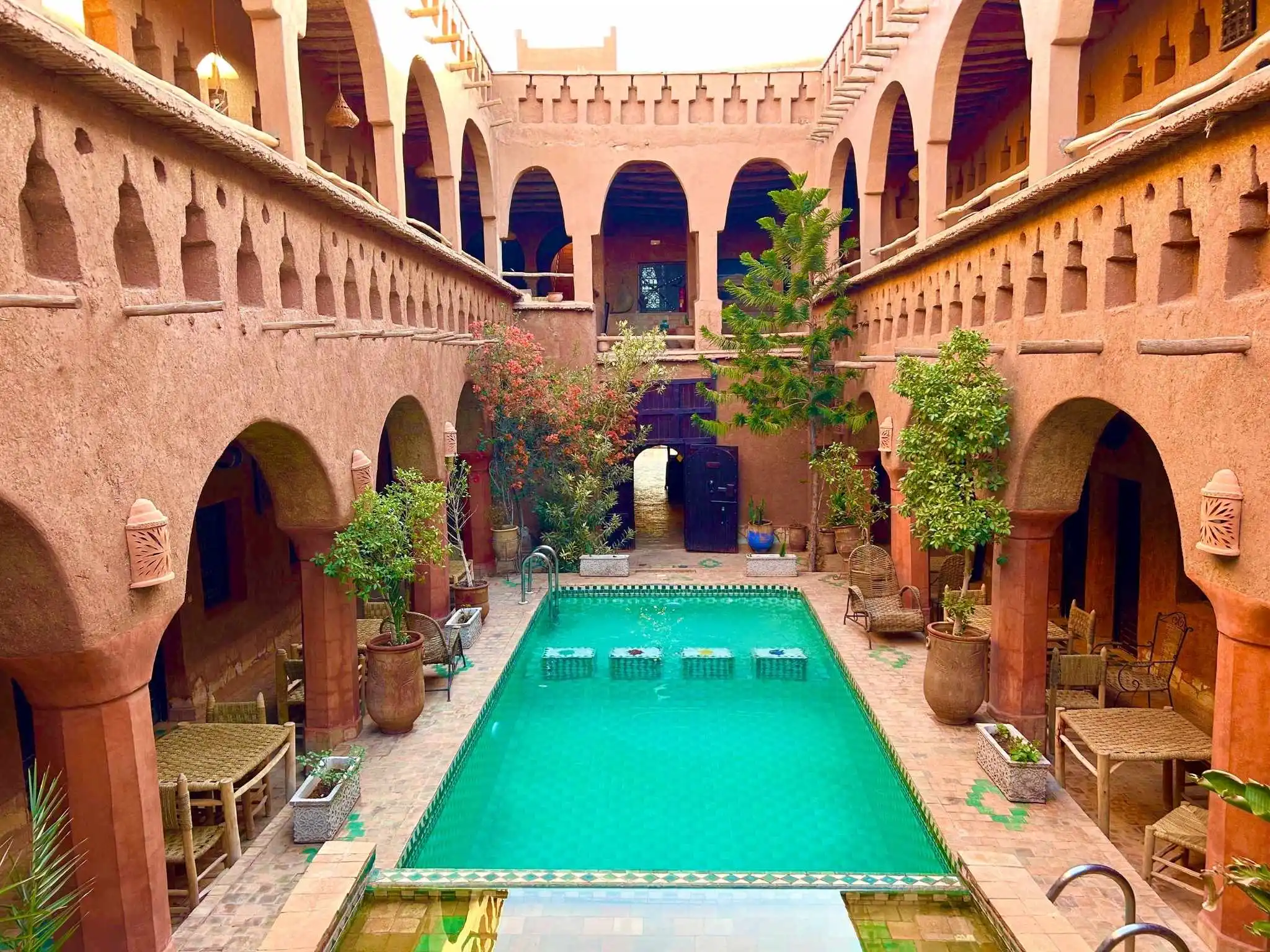 Traditional Moroccan riad with arched galleries and a turquoise tile pool, with tables and potted trees around the courtyard.